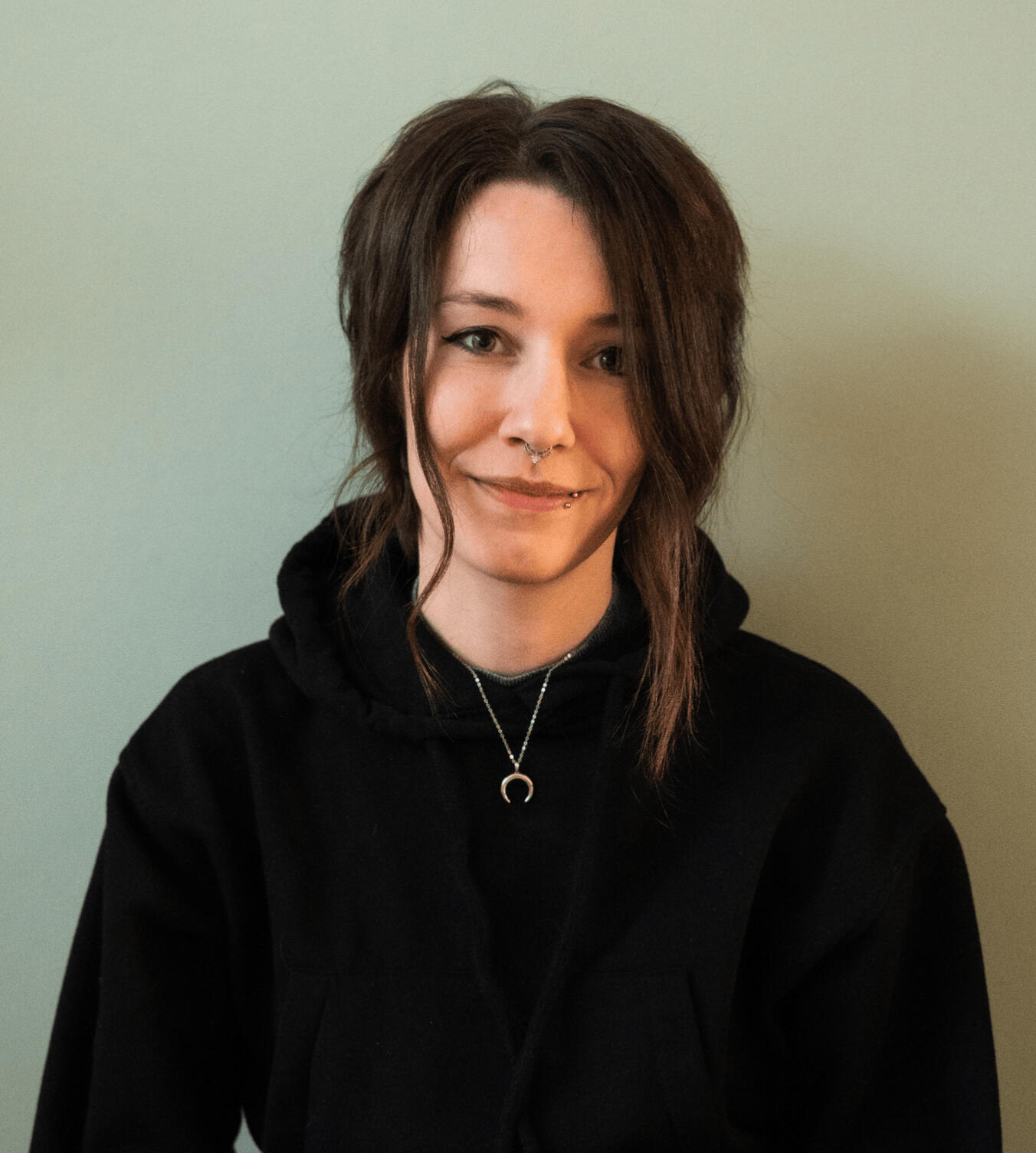 Author portrait of a white person with dark brown hair, cat-eye makeup, a nose and lip piercing, wearing a black sweater.